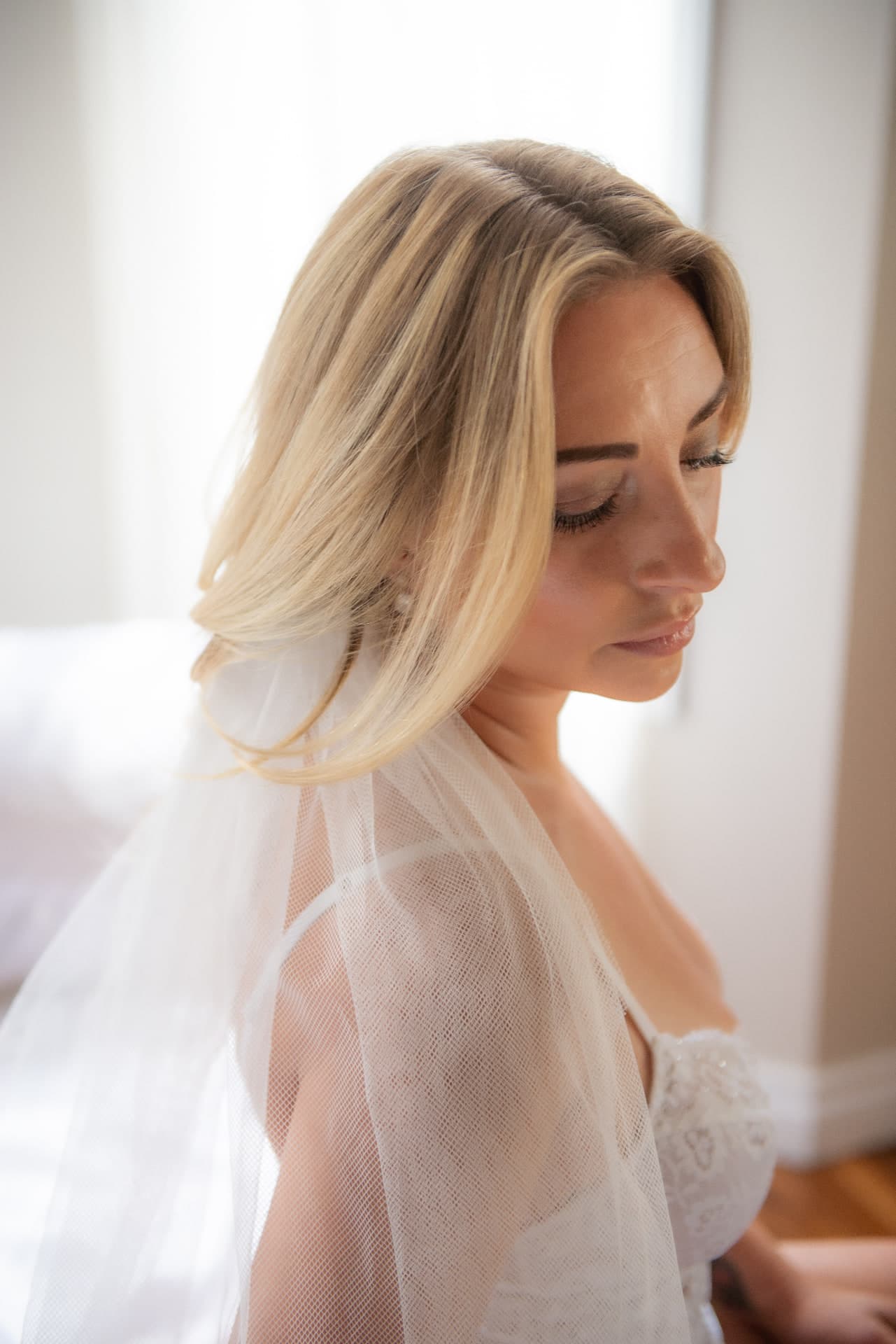 Bride with flowing blonde hair and tulle veil looking down softly, intimate bridal portrait - Bridal Boudoir Edmonton by Touchless Photography